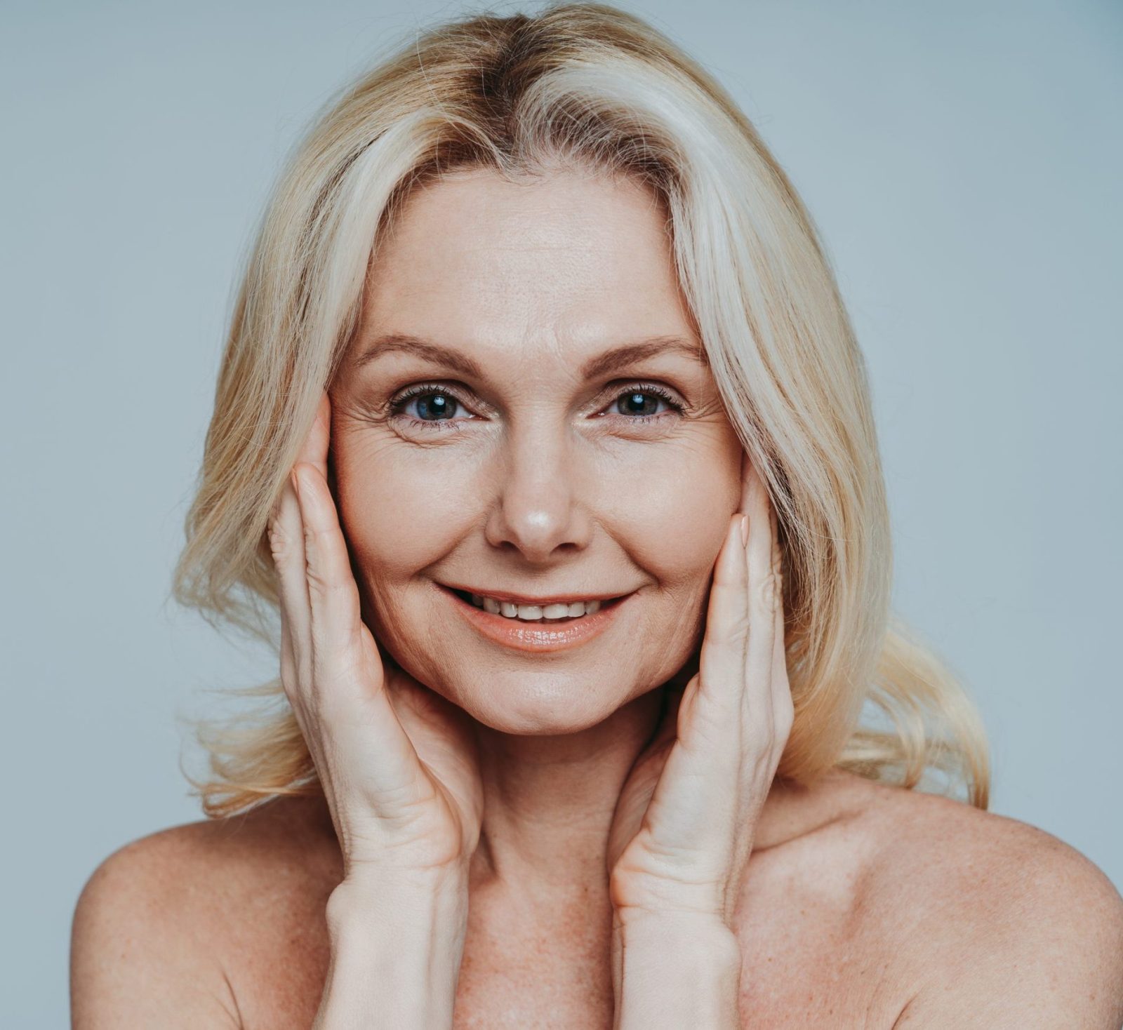 Mature woman with blonde hair smiling at camera, hands framing her face against neutral background